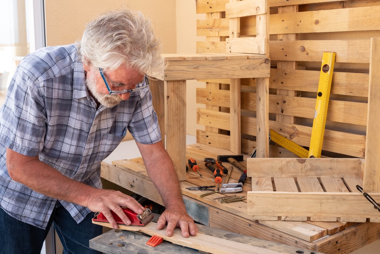 An older man with gray hair and glasses uses a red tool to shape a wooden plank at a workbench, surrounded by wooden pallets, hand tools, and a yellow level.