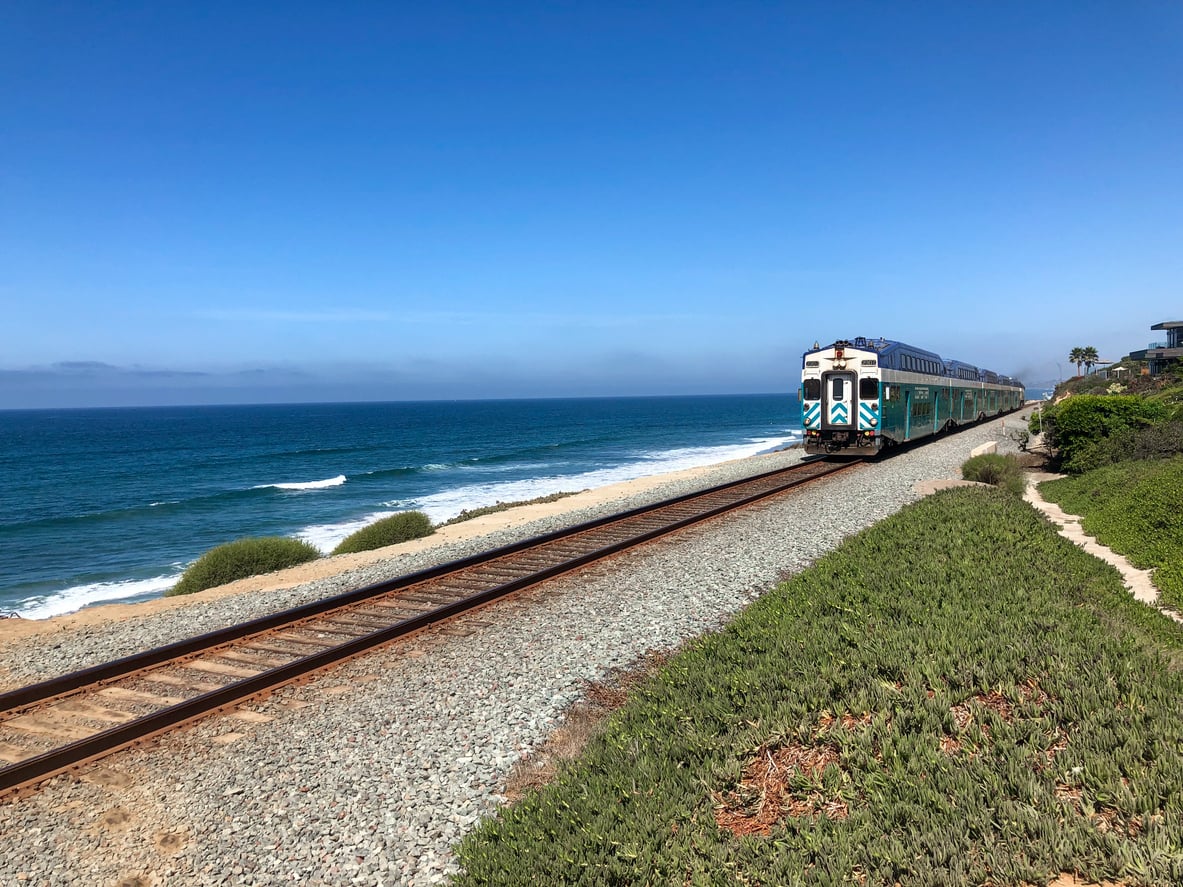 A passenger train travels along coastal railroad tracks next to the ocean, with blue sky above, waves crashing on the shore, and green shrubs lining the tracks.