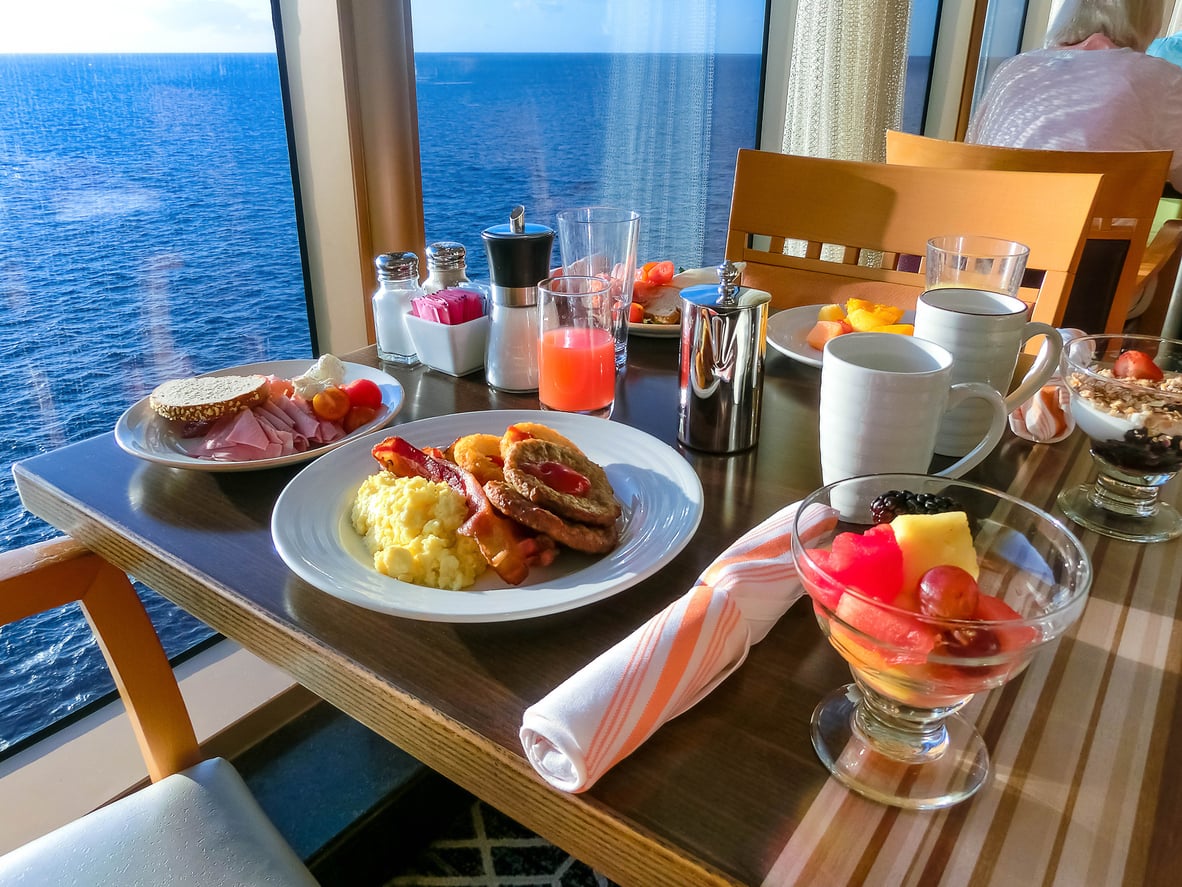 Breakfast spread on a table by a window overlooking the ocean; plates of eggs, bacon, sausage, cold cuts, fruit cups, juice, coffee mugs, and cutlery are arranged on a wooden table with sunlight streaming in.