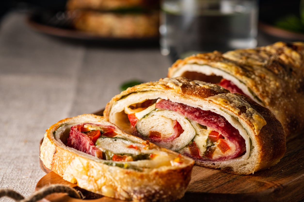 Close-up of a sliced stromboli filled with salami, cheese, and vegetables on a wooden board, with a glass of water and another plate in the blurred background.