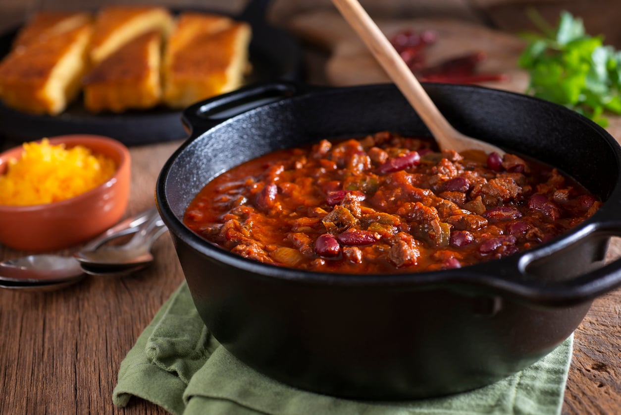 A black pot of chili with beans and meat sits on a green cloth, with a wooden spoon inside. In the background, cornbread and a small bowl of shredded cheese are visible on a wooden table.