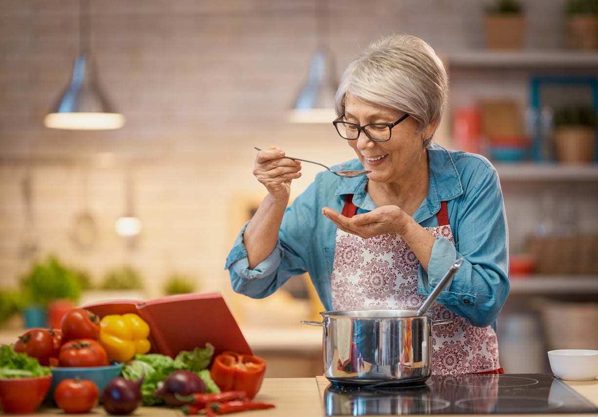 An older woman with gray hair and glasses tastes soup from a spoon in a kitchen. She smiles, wearing an apron, with vegetables and an open cookbook on the counter beside her.
