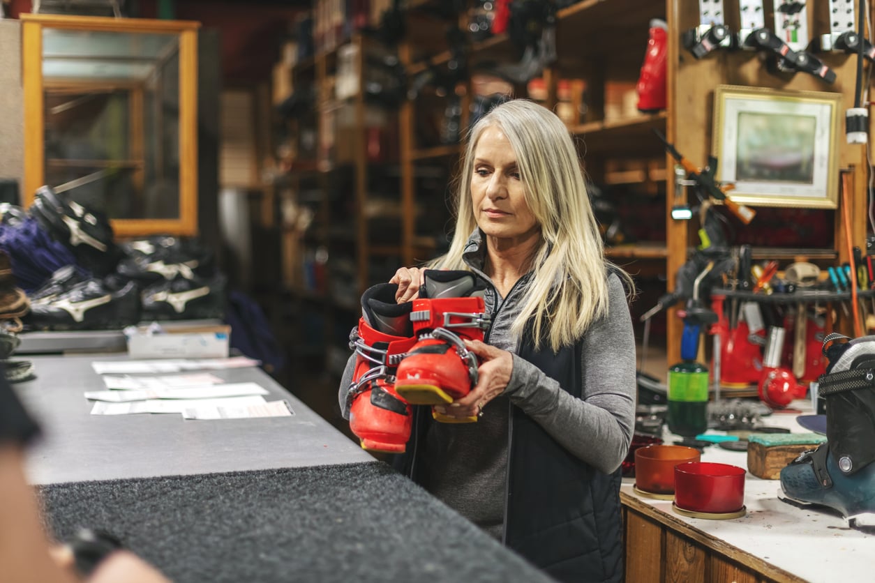 A woman with long gray hair stands behind a counter in a ski shop, holding a pair of red ski boots. Shelves with more ski boots and equipment are visible in the background.