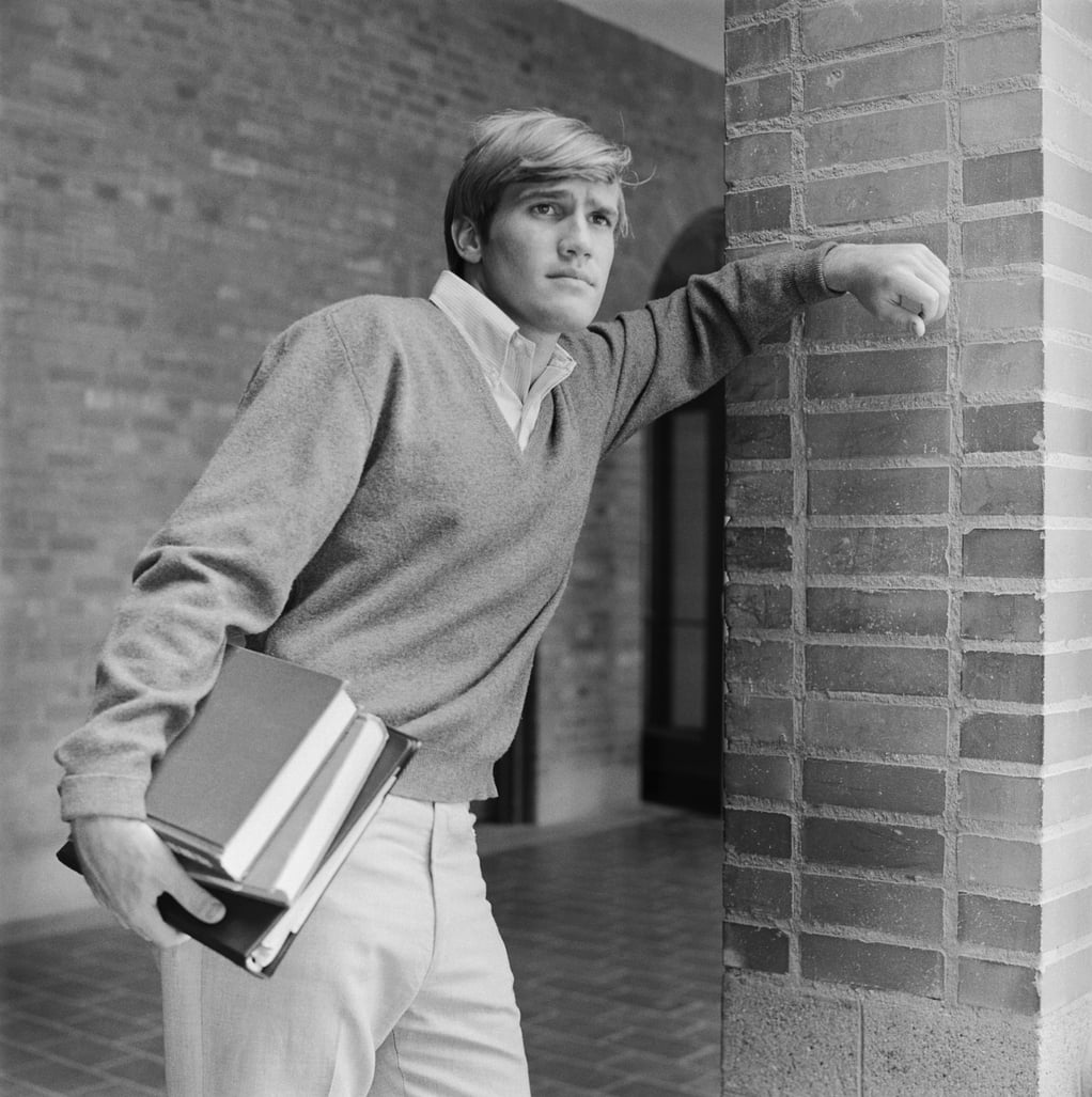 A young man leans against a brick column, holding books in one arm. He wears a sweater over a collared shirt and light pants, looking off into the distance in a hallway with brick walls.
