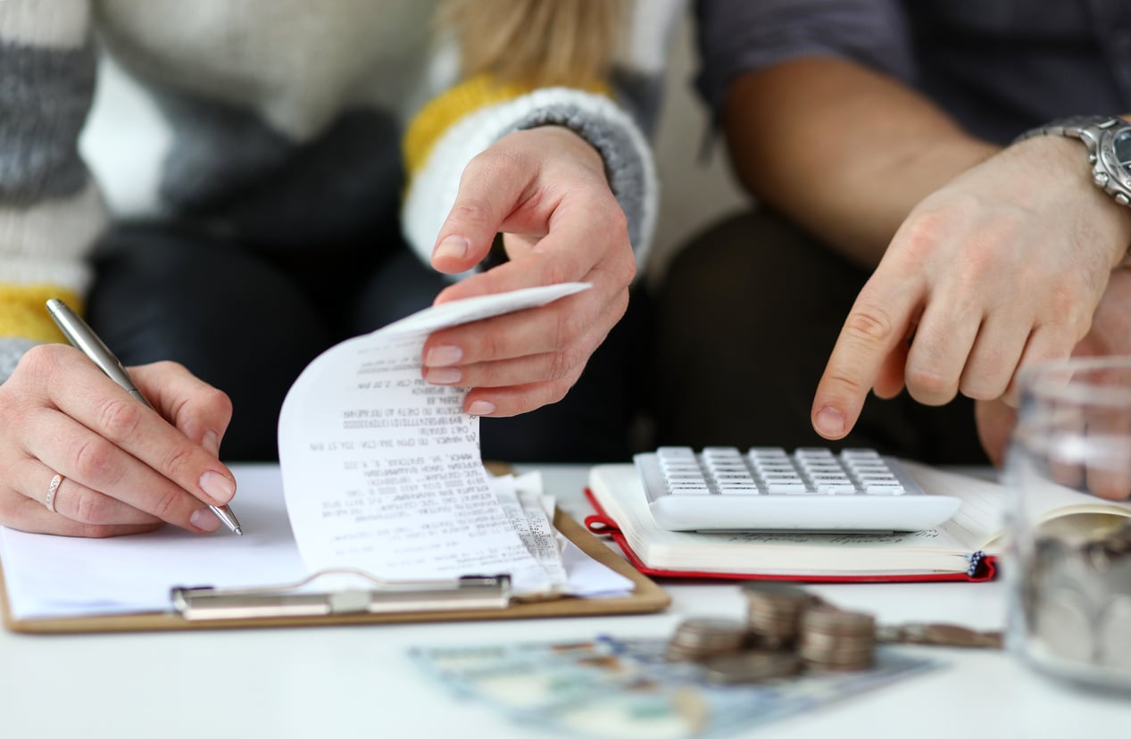 Two people working on finances together, one holding a receipt and writing on a clipboard, the other using a calculator. Coins and paper money are on the table.