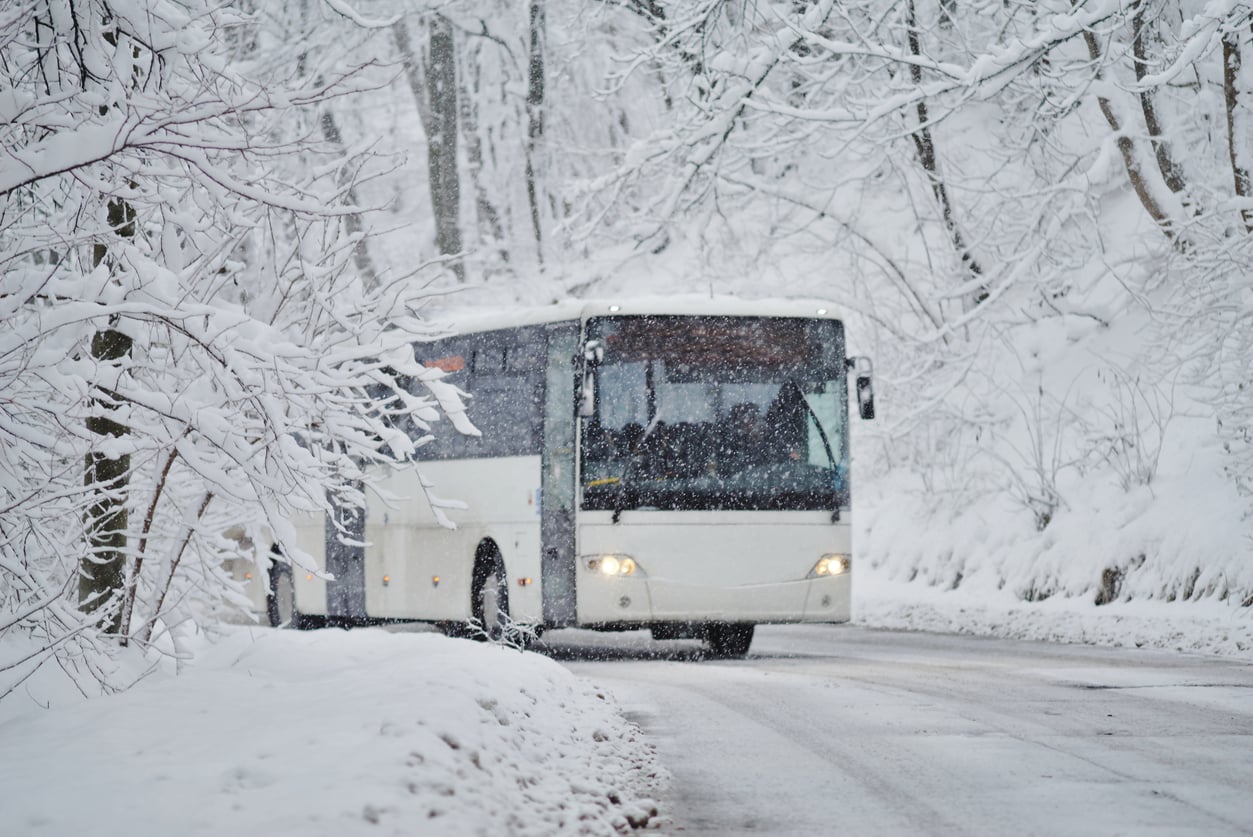 A white bus drives along a snow-covered road surrounded by trees blanketed in heavy snow, creating a wintery scene.