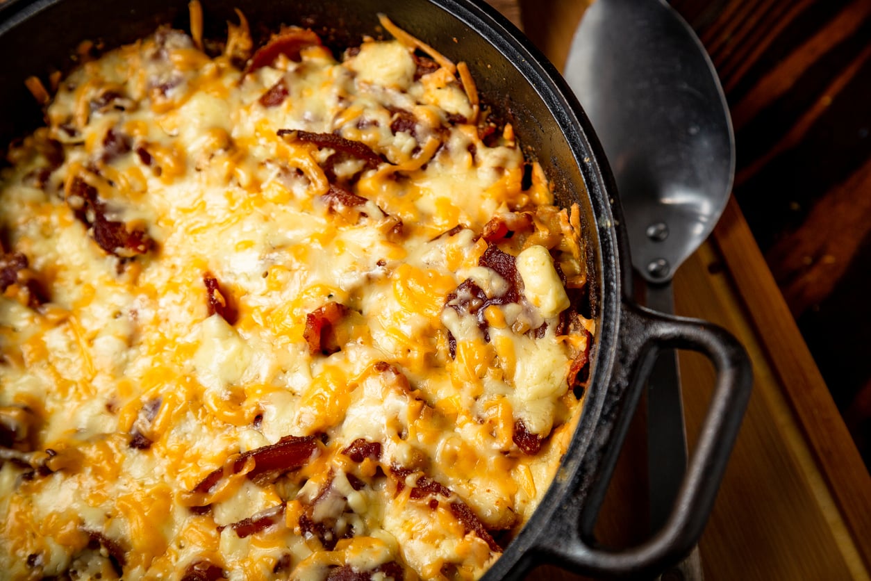 A close-up of a baked casserole in a black skillet, topped with melted cheese and crispy bacon pieces. A metal spatula rests beside the skillet on a wooden surface.