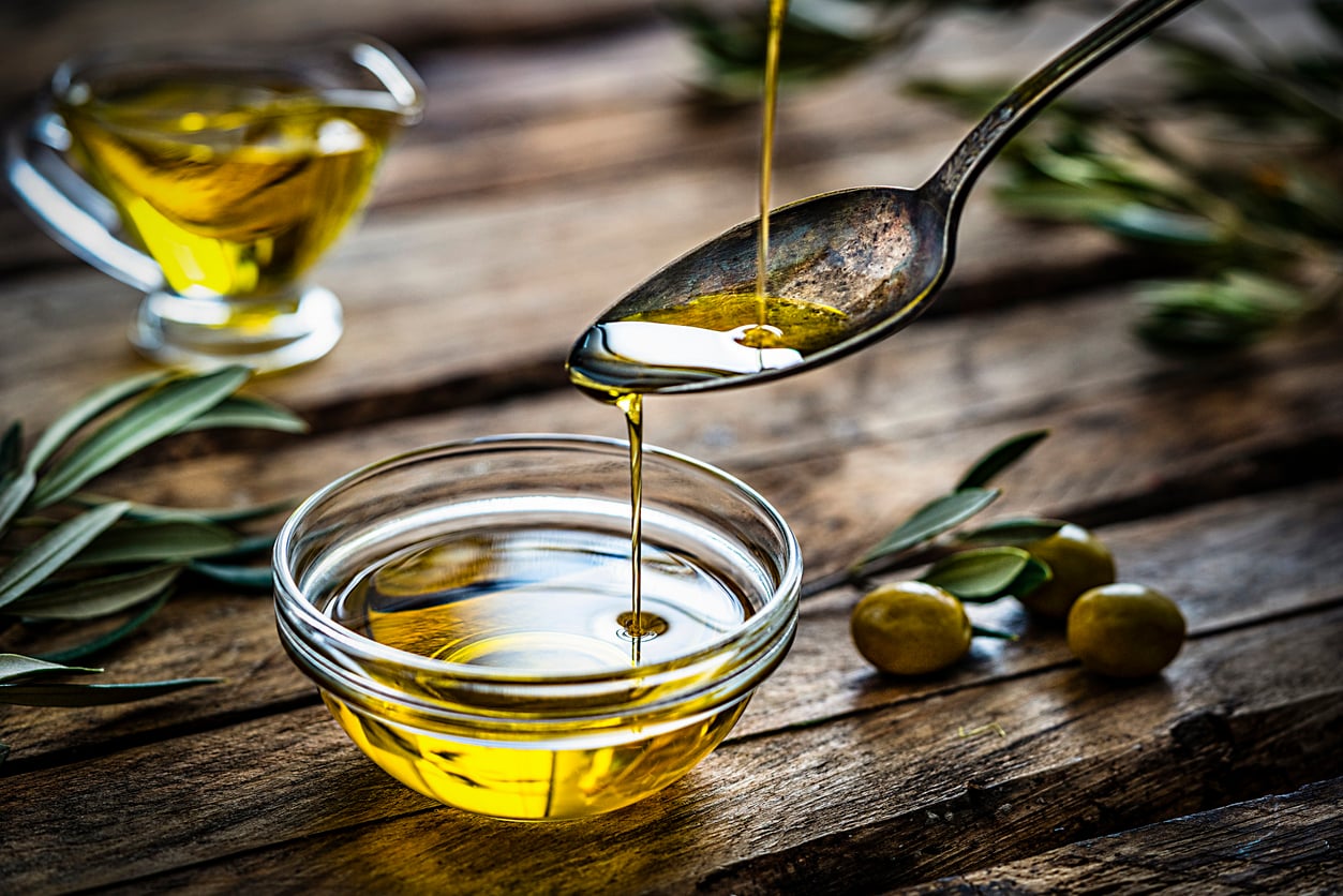 Olive oil being poured from a spoon into a small glass bowl on a rustic wooden table, with olives, leaves, and a glass jug of oil in the background.