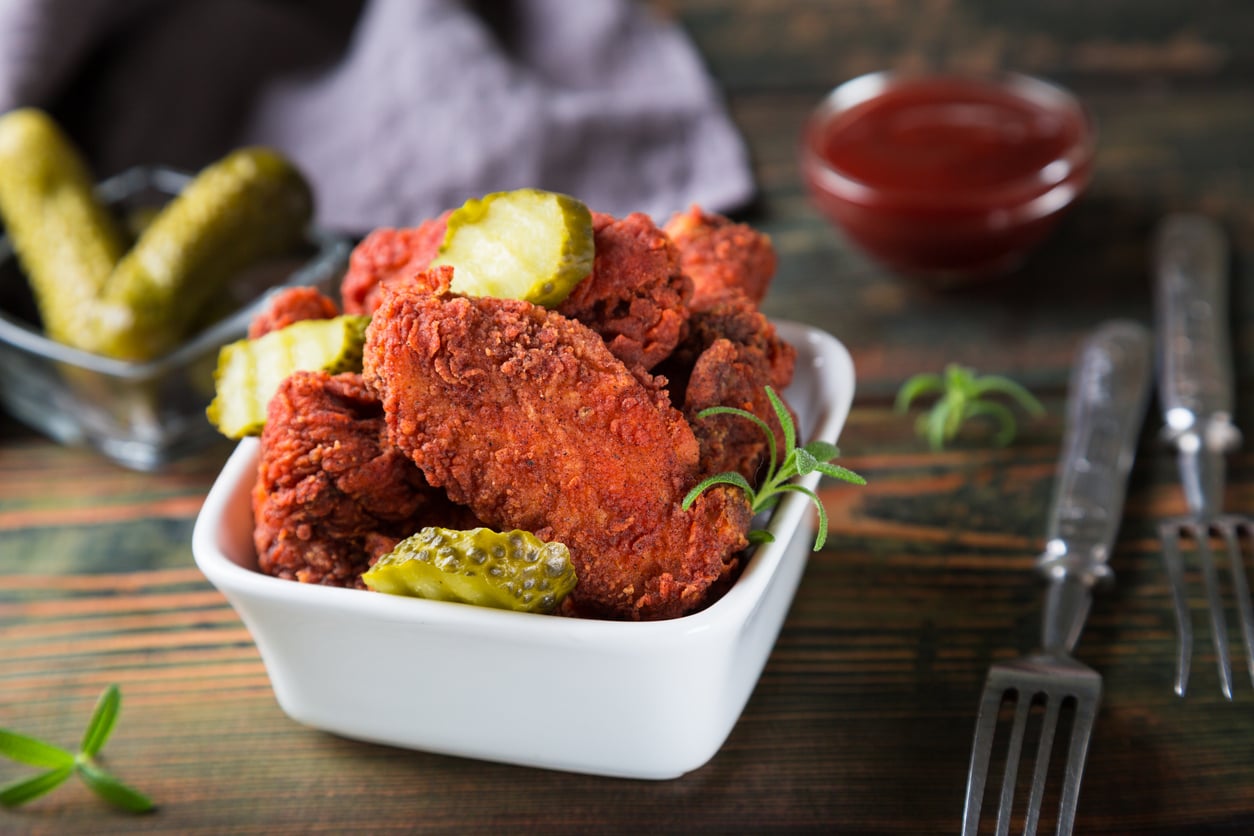 A white bowl filled with crispy fried chicken wings and pickle slices on a wooden table, with a dish of pickles, a cup of sauce, and two forks nearby.