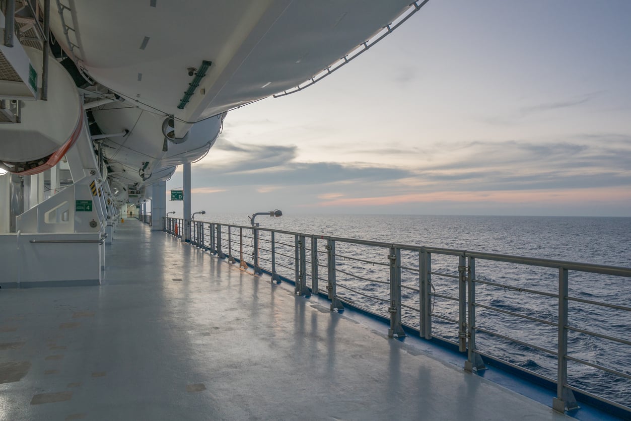 A view from a cruise ship deck at sunset, showing lifeboats above, a metal railing, and calm ocean waters under a cloudy sky.