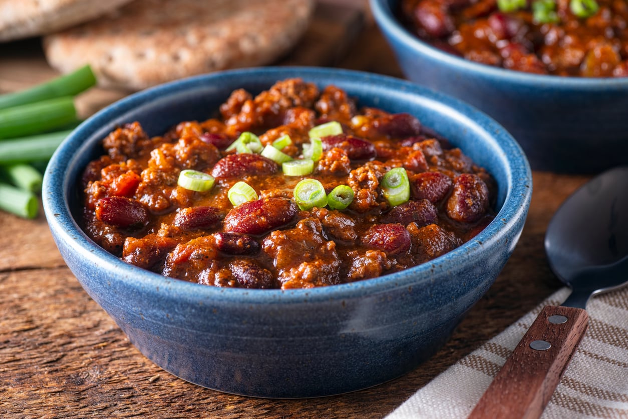 A blue bowl of chili with kidney beans, ground meat, and chopped green onions, featuring hearty foods, is placed on a wooden table next to a spoon, napkin, and some bread in the background.