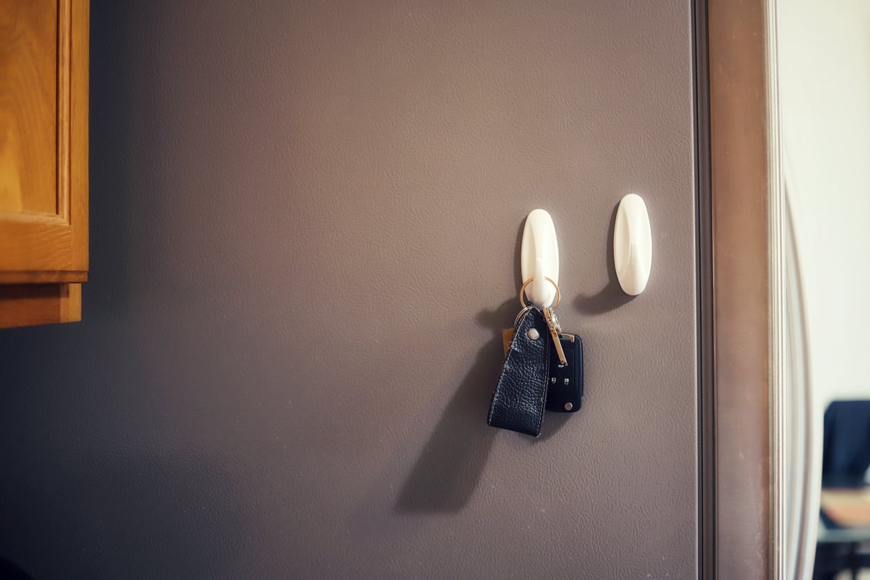 A set of keys with a black leather keychain hangs from a white hook on a gray wall, next to a wooden cabinet.
