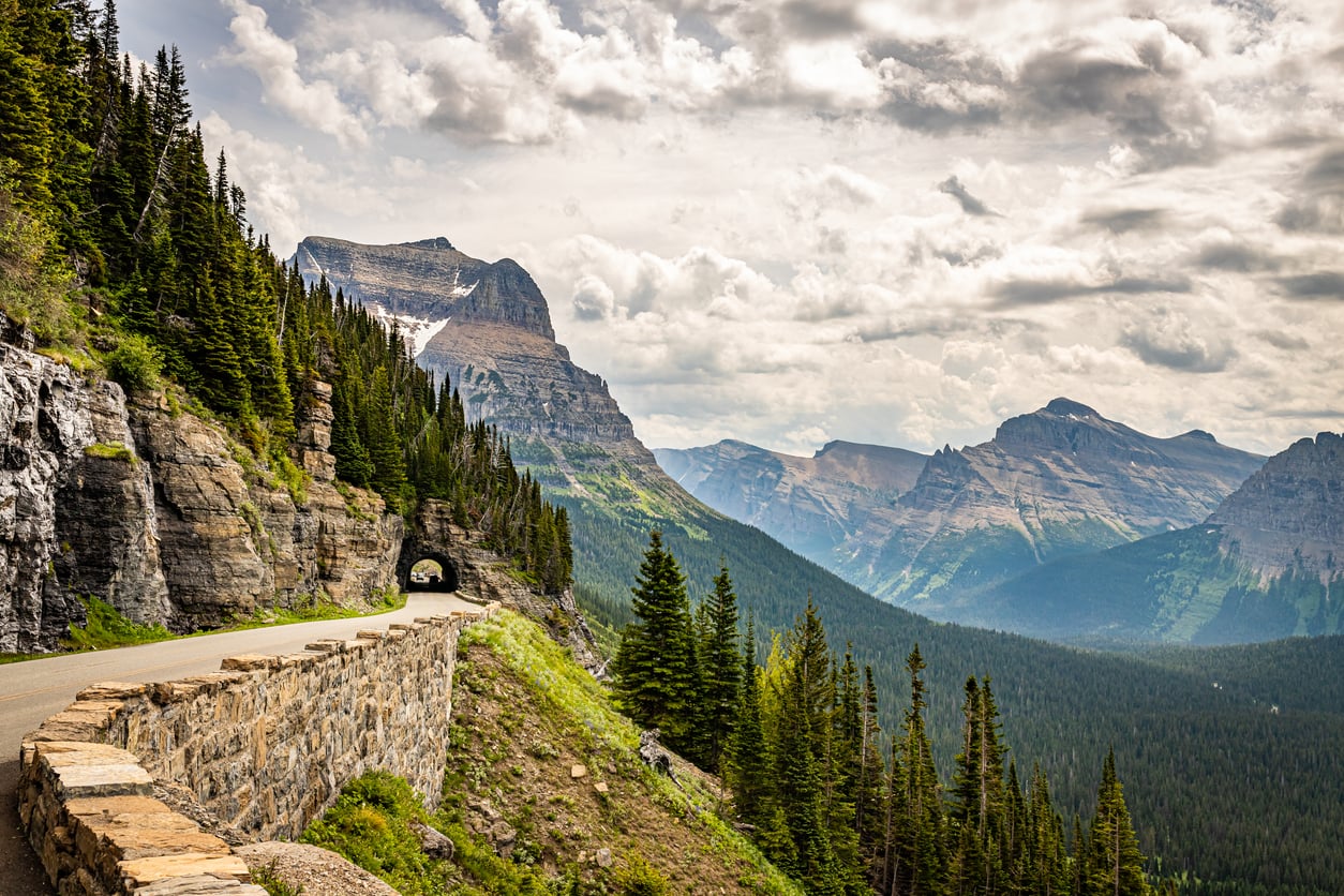 A winding mountain road with a stone barrier curves along a forested slope, passing through a tunnel, with rugged peaks and a partly cloudy sky in the background.