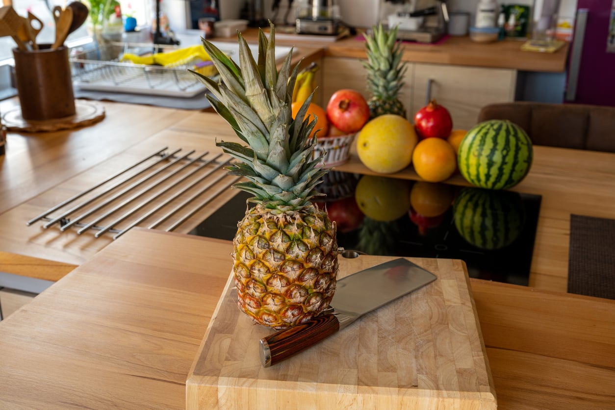 A pineapple and a large knife rest on a wooden cutting board, with various fruits including pomegranate, grapefruit, and watermelon in the background on a modern kitchen counter—an inviting display of everyday foods that used to be considered fancy.