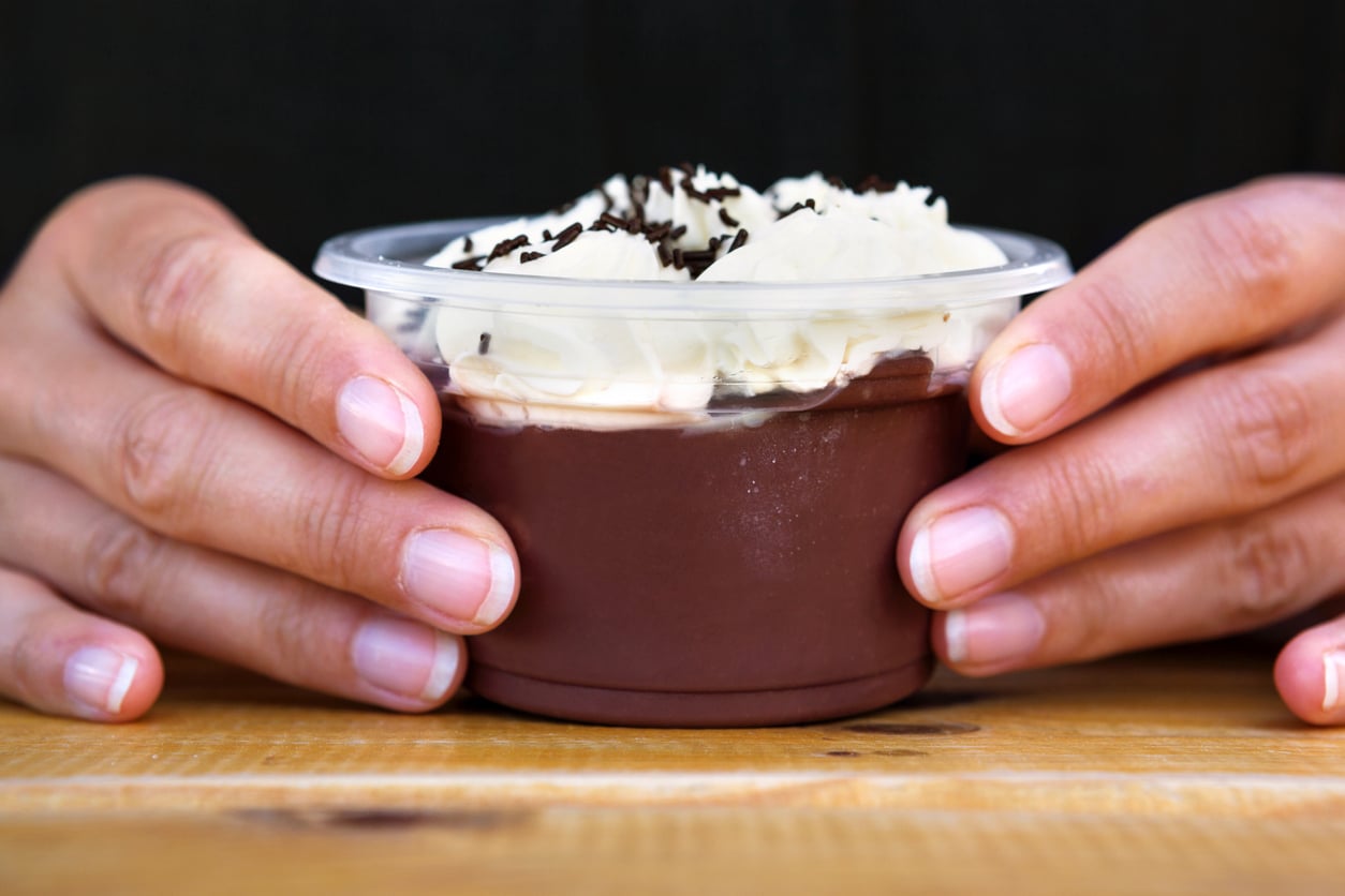 Two hands hold a clear plastic cup filled with chocolate pudding, topped with whipped cream and chocolate sprinkles, on a wooden table.
