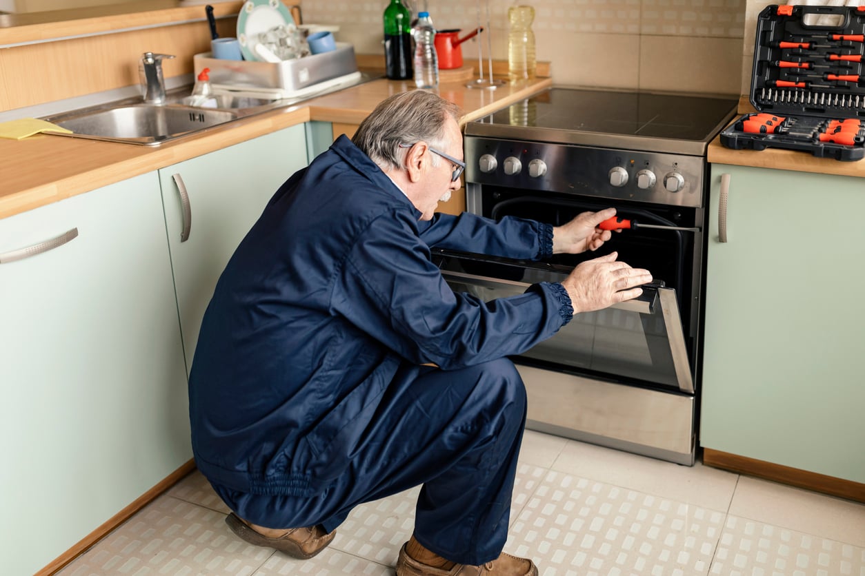 An older man, one of the Baby boomers, in blue coveralls repairs an oven in a kitchen, holding a screwdriver while examining the oven door. Tools and kitchen items are visible around him.