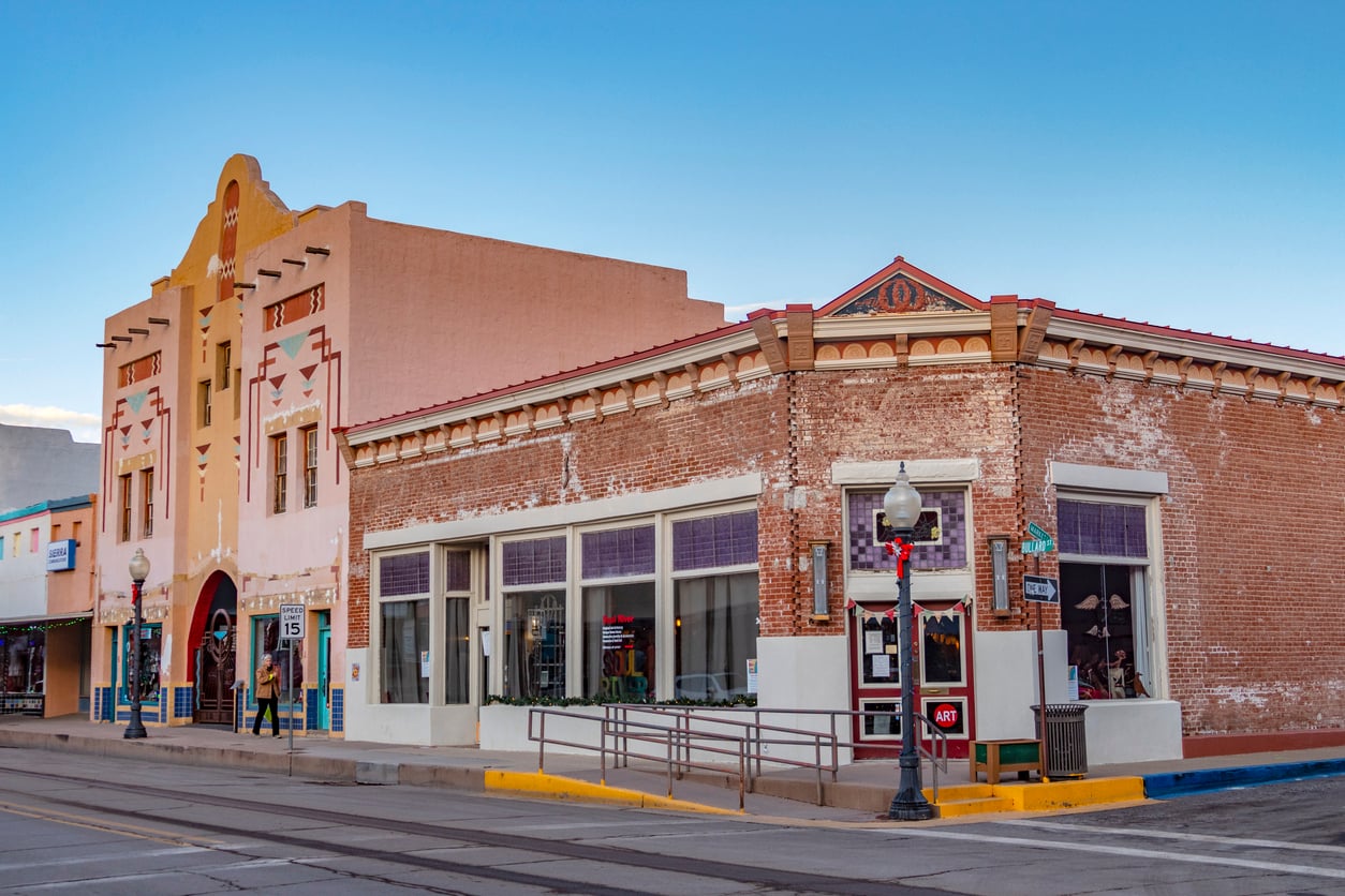 A street view of two historic buildings, one with an Art Deco facade and the other with exposed brick. Both have storefront windows and vintage street lamps line the sidewalk. The sky is clear and blue.