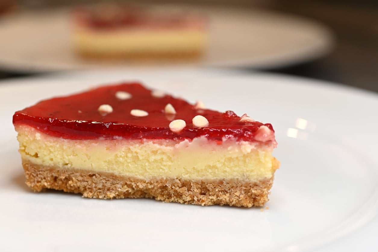 A slice of cheesecake with a biscuit crust, creamy filling, and a glossy red fruit topping, decorated with small white sprinkles, served on a white plate.