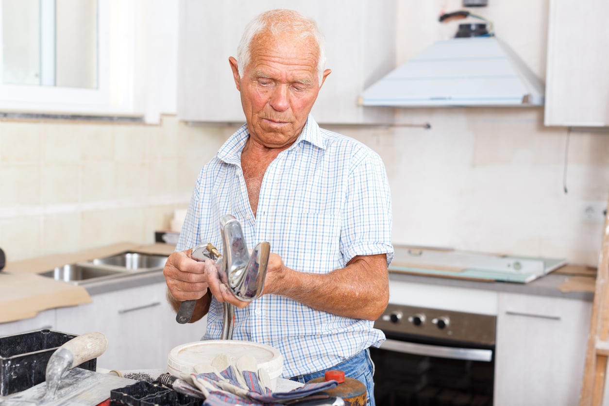 An elderly man in a plaid shirt repairs a kitchen faucet in a modern kitchen, holding tools and looking focused on his work.
