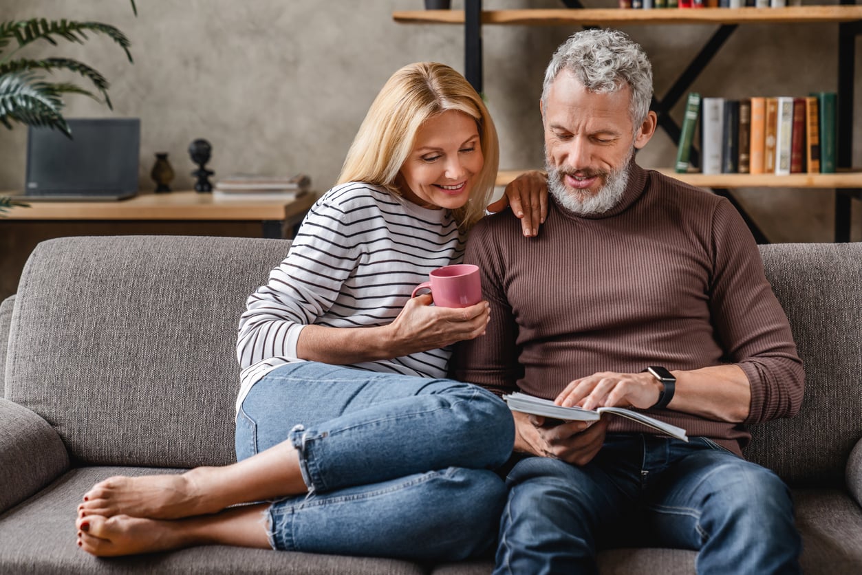 A smiling couple of adults over 50 sits on a couch; the woman holds a pink mug while the man shows her something in an open book. They look relaxed and comfortable in a cozy living room with a bookshelf in the background.
