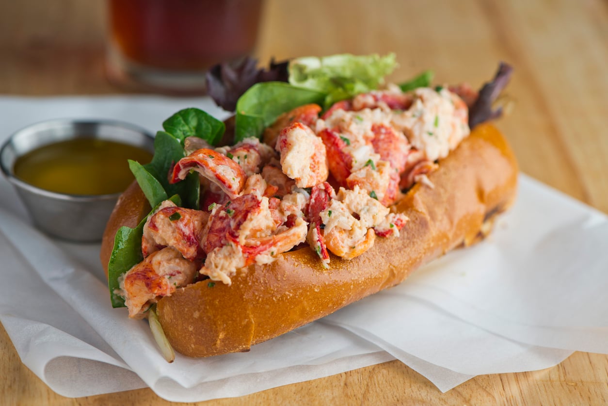 A lobster roll on a toasted bun with leafy greens, served on parchment paper with a side of melted butter and a drink in the background.
