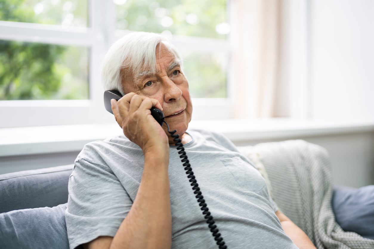 Elderly man with white hair sits on a couch indoors, holding a corded phone to his ear and looking thoughtful. Light streams in through a window behind him.