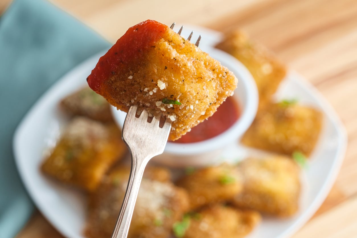 A fork holds a breaded, fried ravioli dipped in marinara sauce, with more ravioli and a bowl of marinara on a white plate in the background.