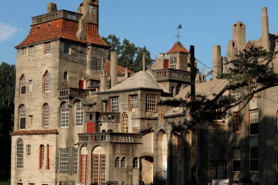 An eclectic concrete castle with red-framed windows, multiple turrets, and unique rooflines evokes the timeless charm of classic castles, surrounded by trees and bathed in warm sunlight.