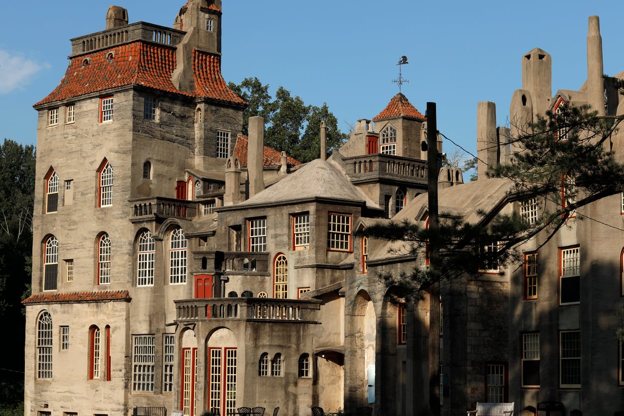 An eclectic concrete castle with red-framed windows, multiple turrets, and unique rooflines evokes the timeless charm of classic castles, surrounded by trees and bathed in warm sunlight.