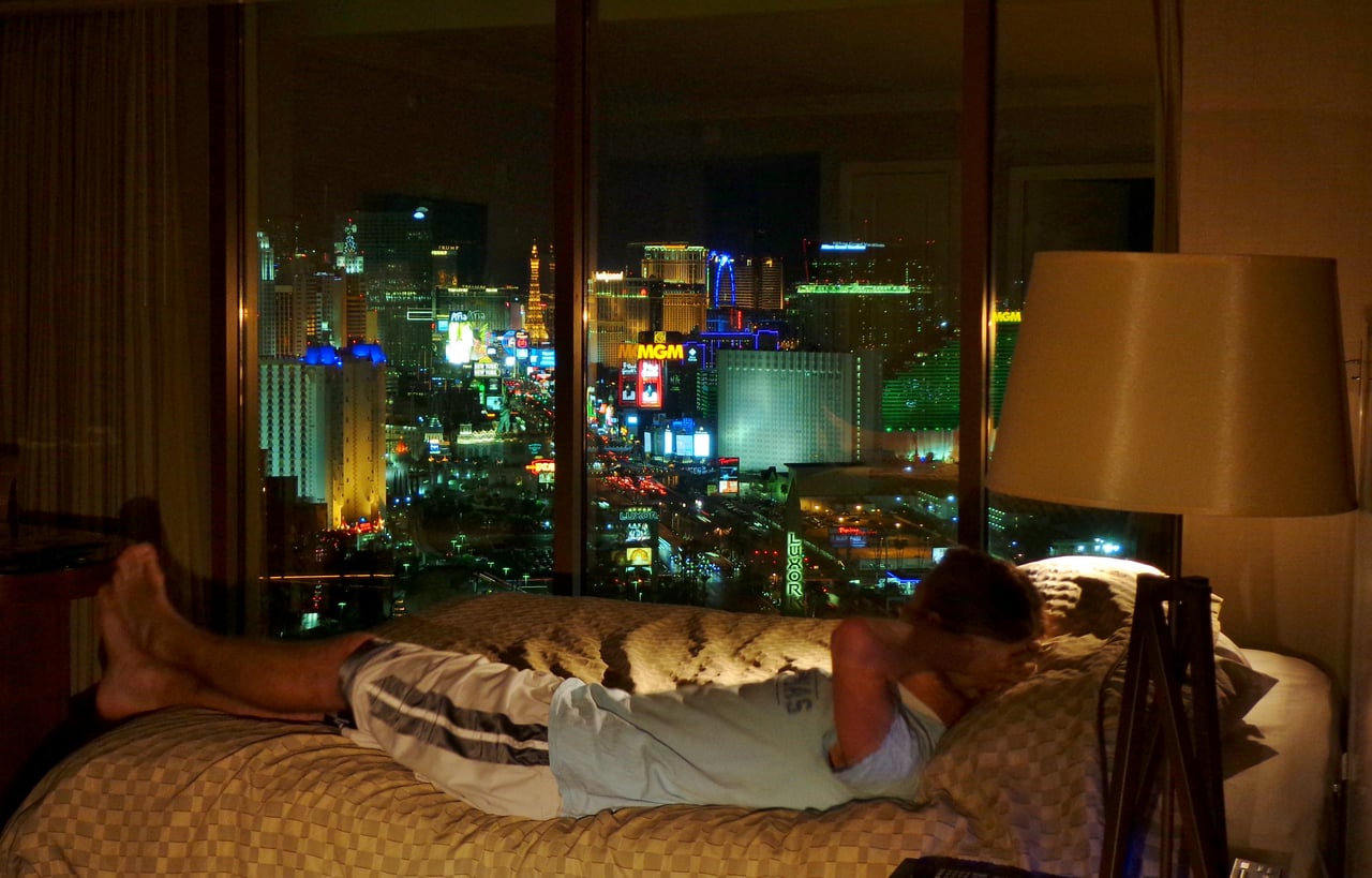 A person lies on a bed in a dimly lit hotel room at night, with a large window showing a vibrant cityscape of bright lights and buildings outside.
