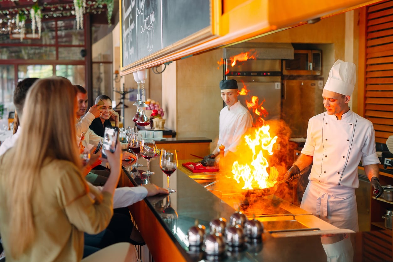 A chef in a white uniform cooks with a burst of flames on a teppanyaki grill as diners at a counter watch and take photos; wine glasses are on the counter.