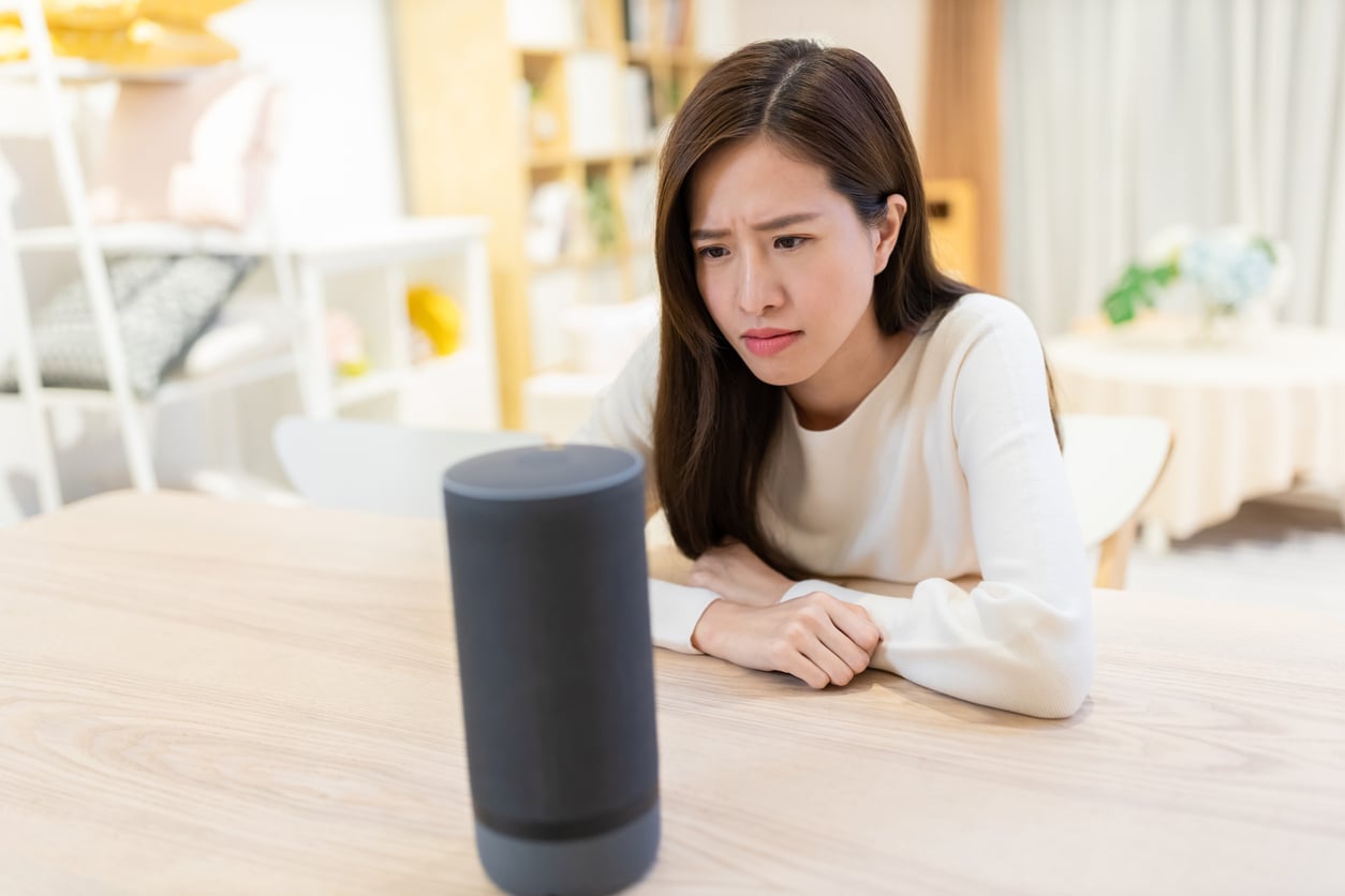 A woman sits at a table, looking confused or frustrated at a smart speaker device in front of her.
