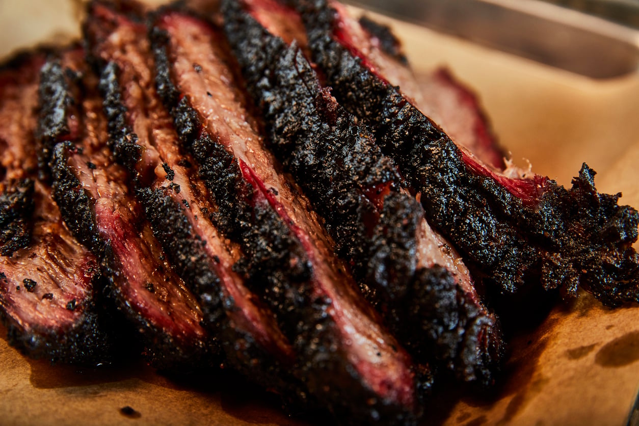 Several slices of smoked brisket with a dark, crispy bark and juicy, tender interior are arranged on brown parchment paper.