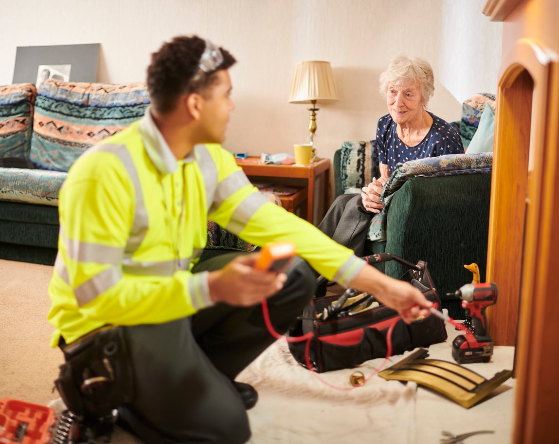 A utility worker in a high-visibility jacket kneels on the floor with tools, checking wires, while an elderly woman sits on a couch watching him inside a living room.