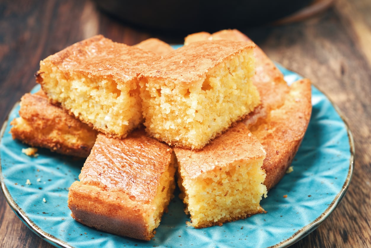 Six golden, moist pieces of cornbread—one of the most comforting foods—are stacked on a blue patterned plate, sitting on a rustic wooden table. The cornbread has a crisp edge and a soft, crumbly texture.