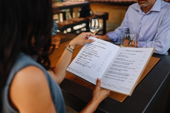 A woman holds a menu at a restaurant table, seated across from a man in a light blue shirt. Both have empty wine glasses in front of them, and the table is set for dining.