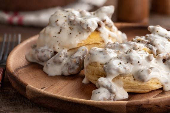 Two flaky biscuits topped with creamy sausage gravy and black pepper, served on a wooden plate, make for one of the most comforting foods. A fork is visible on the side, and the background has a rustic, cozy kitchen feel.