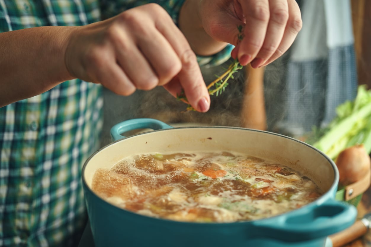 A person in a plaid shirt adds fresh herbs to a pot of simmering soup filled with vegetables.