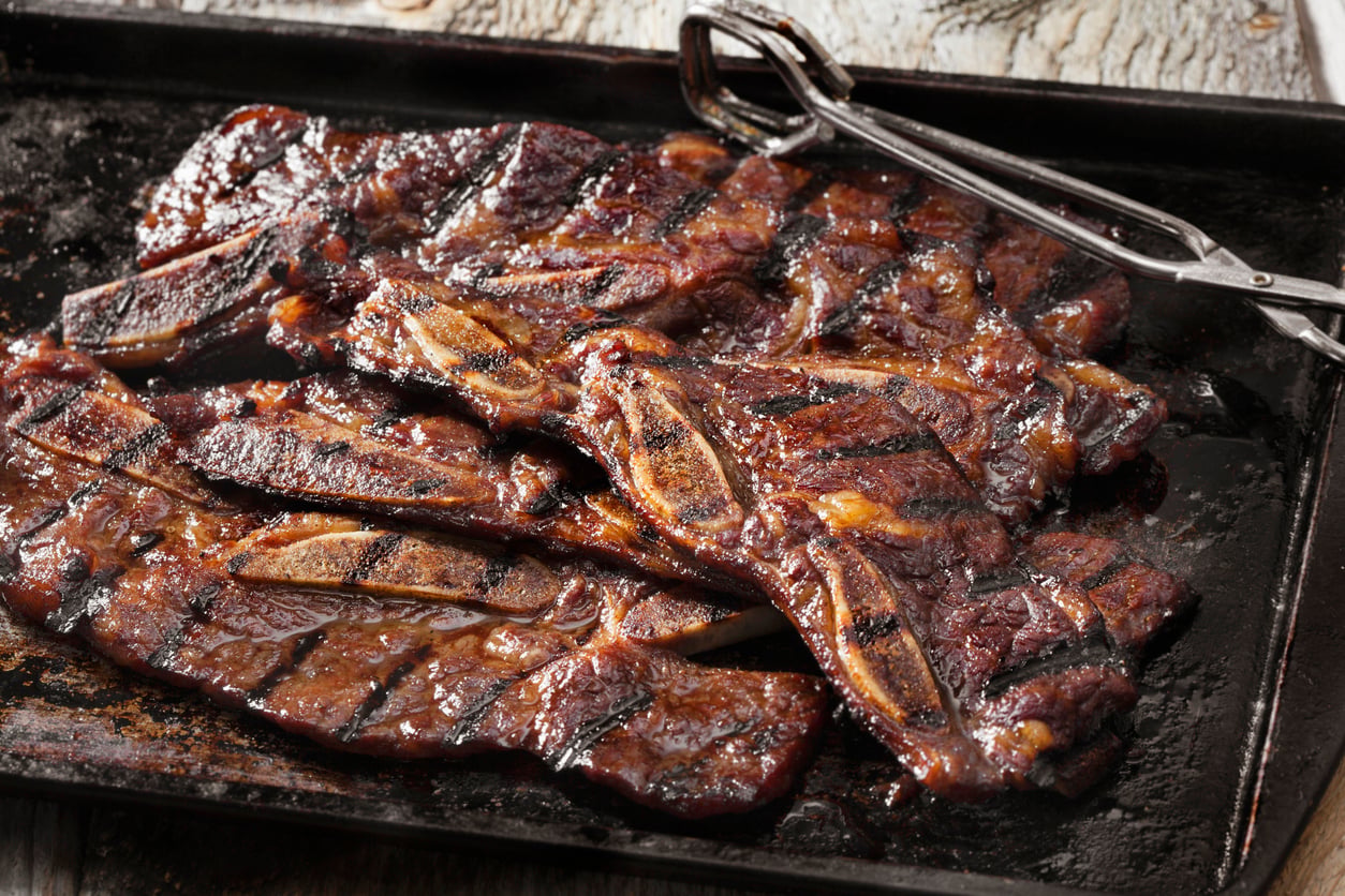 Grilled marinated beef short ribs with visible grill marks are arranged on a dark tray. Metal tongs rest beside the meat. The surface looks juicy and caramelized, with a rustic wooden table underneath.