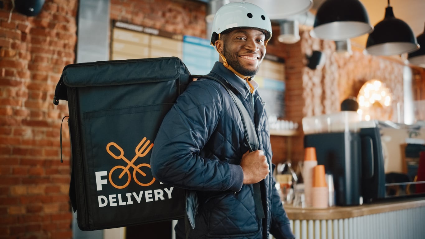 A smiling food delivery worker wearing a helmet and a blue jacket stands indoors, carrying a large insulated delivery bag with “FOOD DELIVERY” written on it. The background shows a café with brick walls and hanging lights.