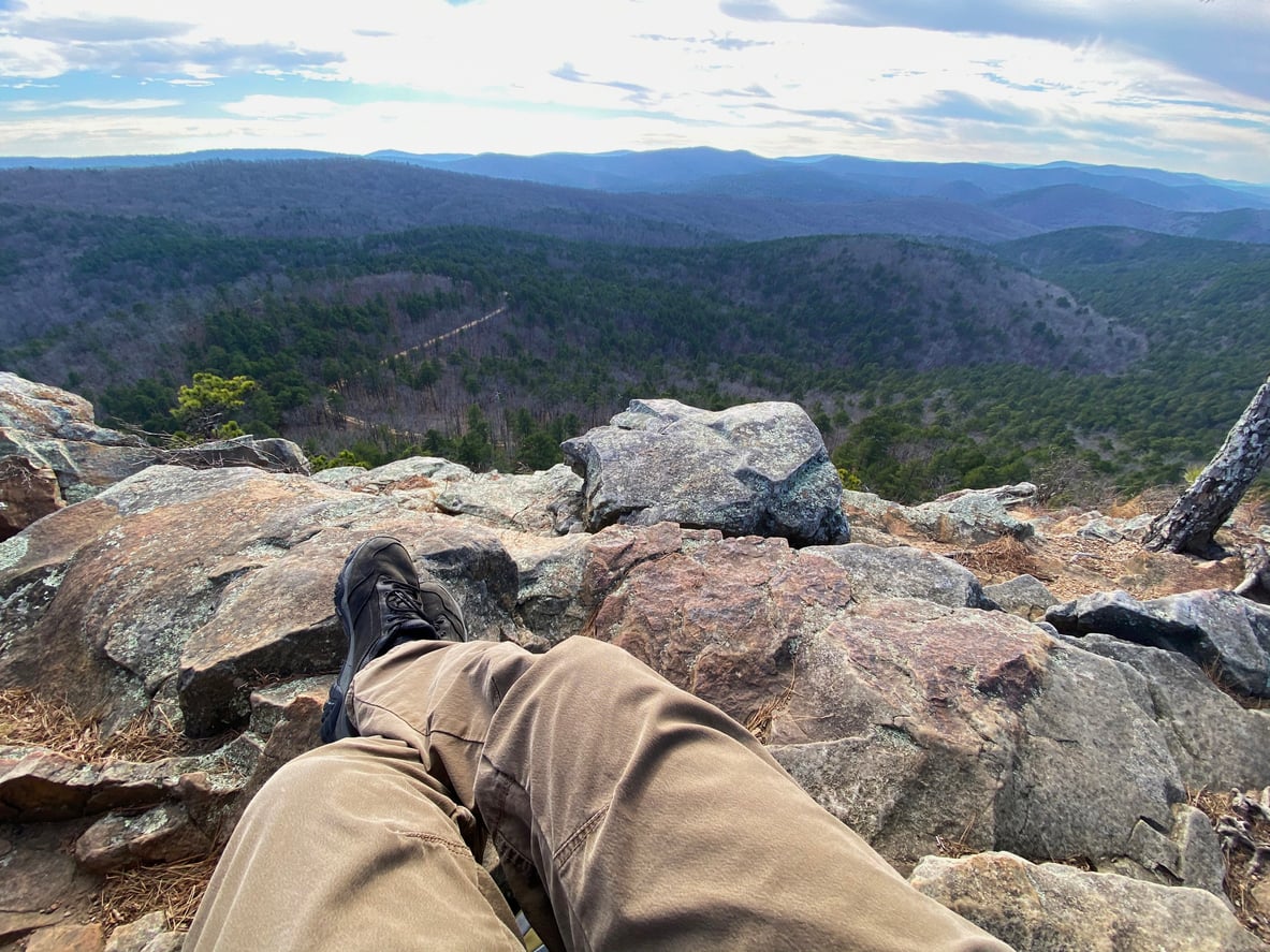 Person in tan pants and black boots sitting on rocky ground, overlooking a vast, tree-covered mountain landscape—one of the region’s most breathtaking hiking destinations—under a partly cloudy sky.