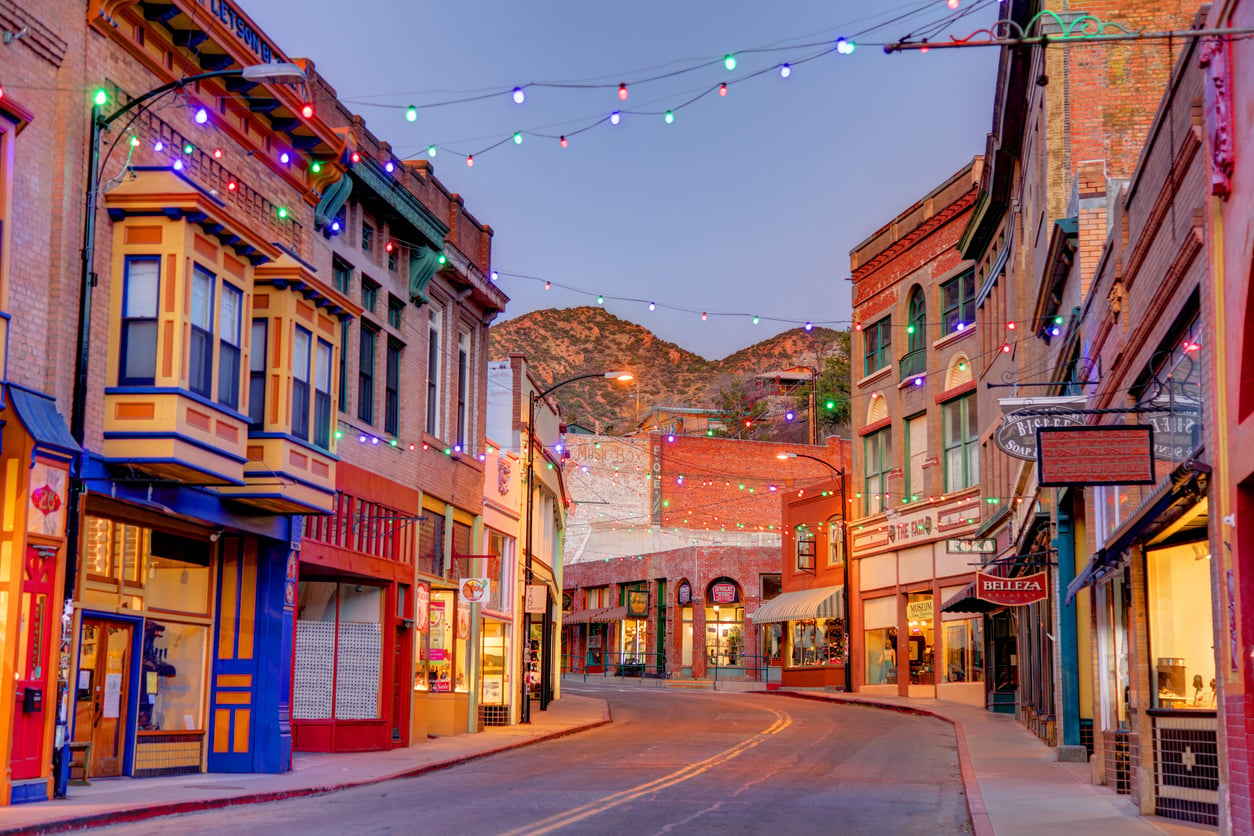 A colorful, empty street in a small town with historic brick buildings, string lights overhead, and mountains in the background at dusk.