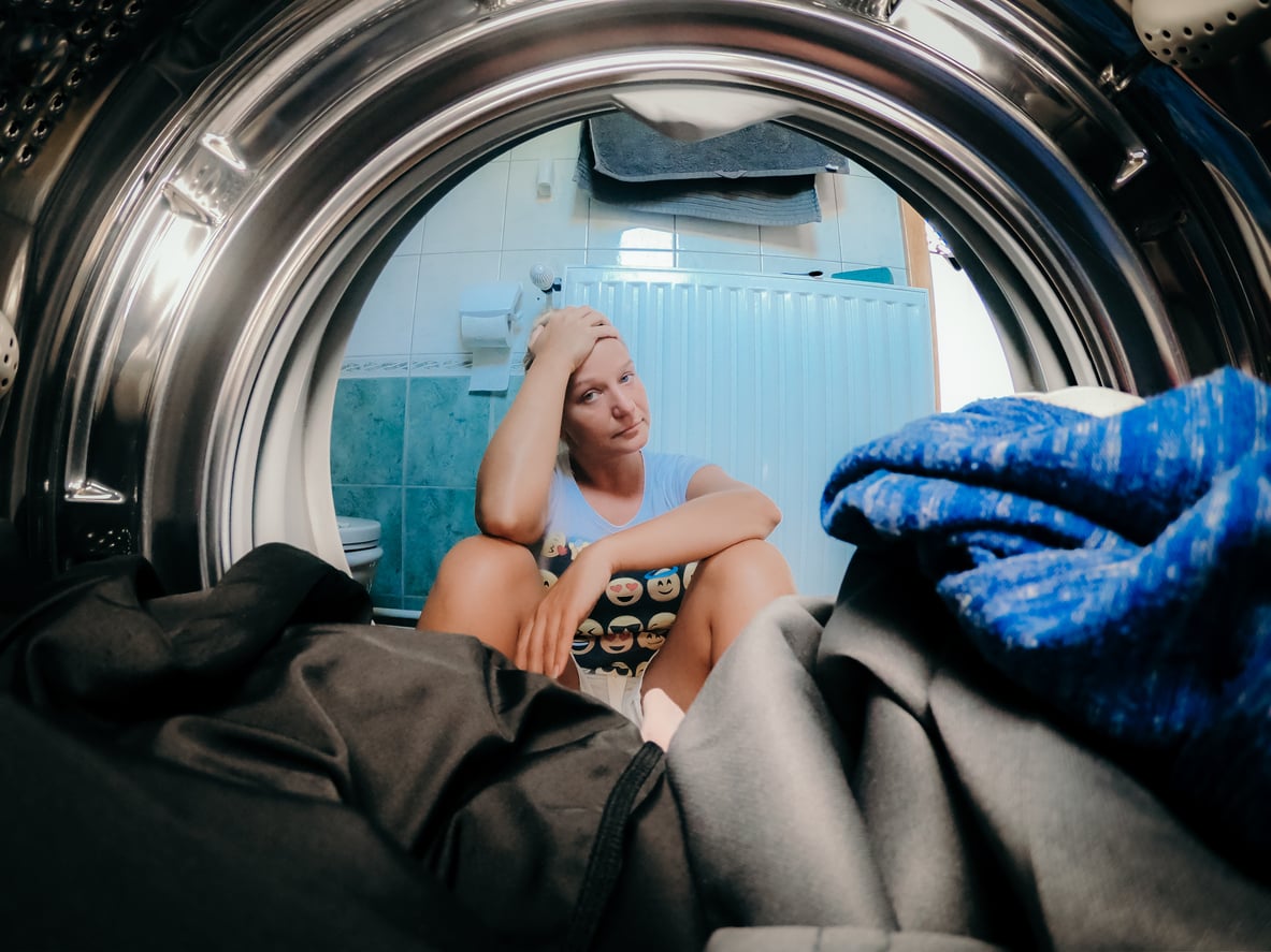 A person sits on the bathroom floor, looking tired and holding their head, as seen from inside a washing machine filled with laundry.