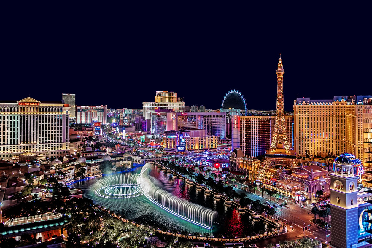 Las Vegas Strip at night, featuring brightly lit hotels, casinos, the Eiffel Tower replica, and the Bellagio fountains with water jets and circular patterns illuminated against the dark sky.