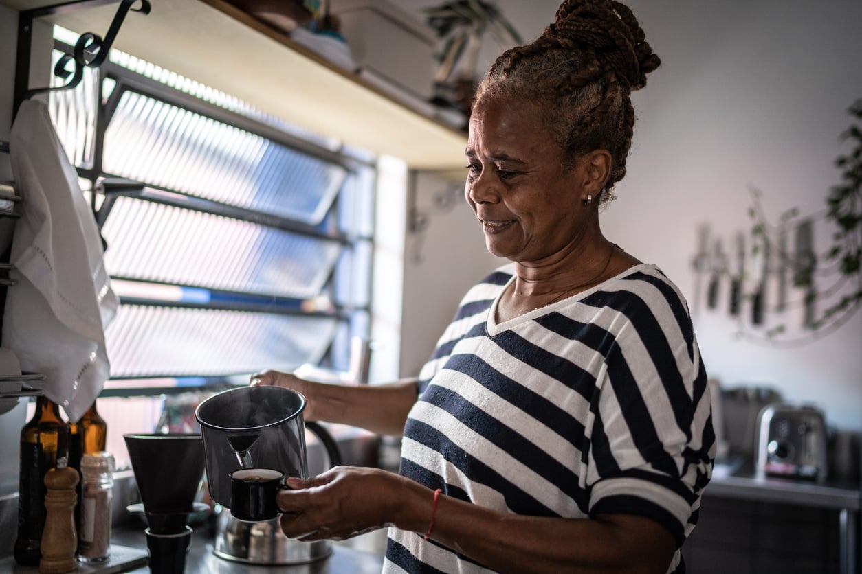 A woman in a striped shirt smiles while pouring coffee from a kettle into a cup in a bright kitchen with plants and kitchenware in the background.