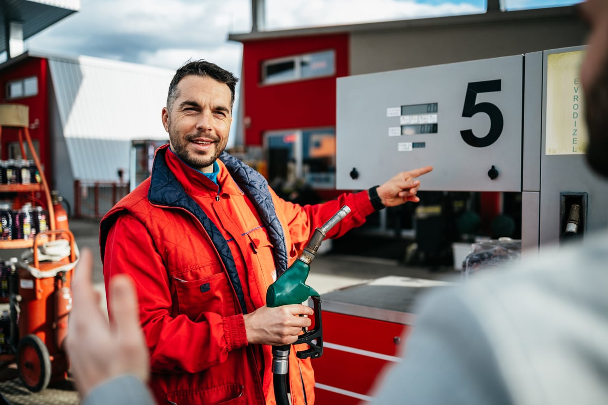 A man in a red jacket, possibly on vacation, holds a fuel nozzle and points at a gas pump numbered 5 at a gas station, interacting with someone off-camera.