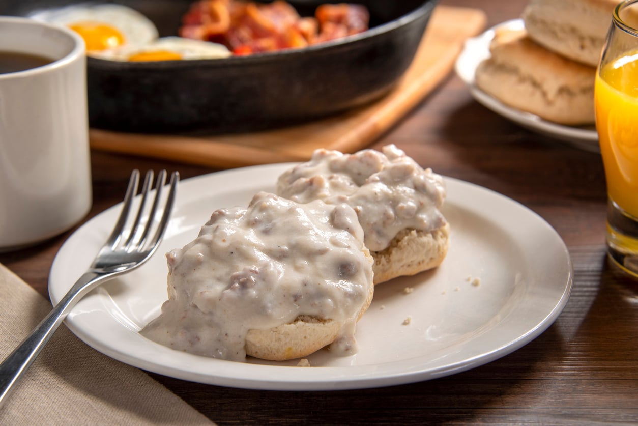 A plate with two biscuits covered in sausage gravy sits next to a fork. In the background are eggs, bacon in a skillet, biscuits, a cup of coffee, and a glass of orange juice.