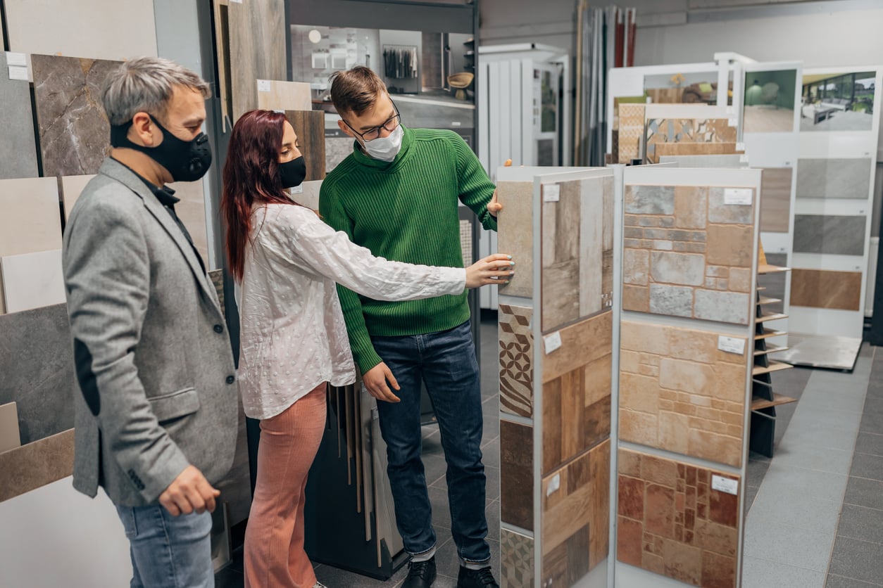 Three people wearing face masks look at various tile samples in a store. One person points to a display while the others observe, suggesting they are discussing or choosing tiles for a project.