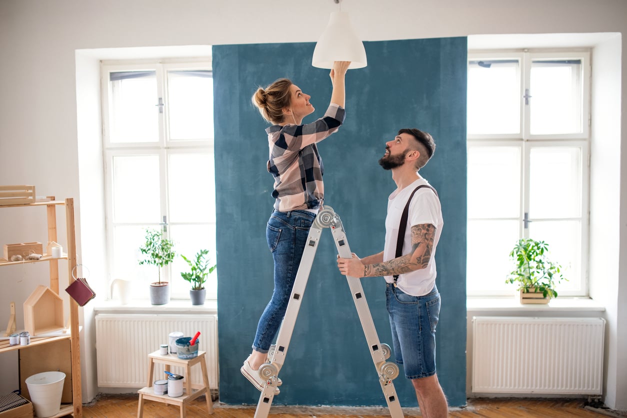 A woman stands on a ladder adjusting a ceiling light while a man holds the ladder. They are in a bright room with large windows, houseplants, and a blue-painted wall.