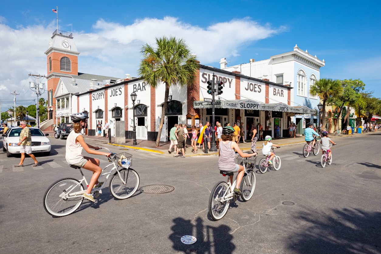 People ride bikes past Sloppy Joe’s Bar on a sunny day as pedestrians walk by; palm trees and historic buildings line the street in a lively Key West scene.
